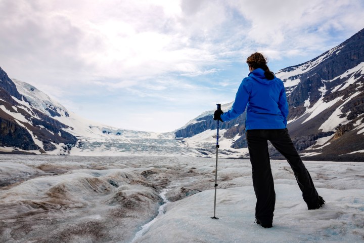 Person in blue jacket hiking on glacier with snowy mountains in background.