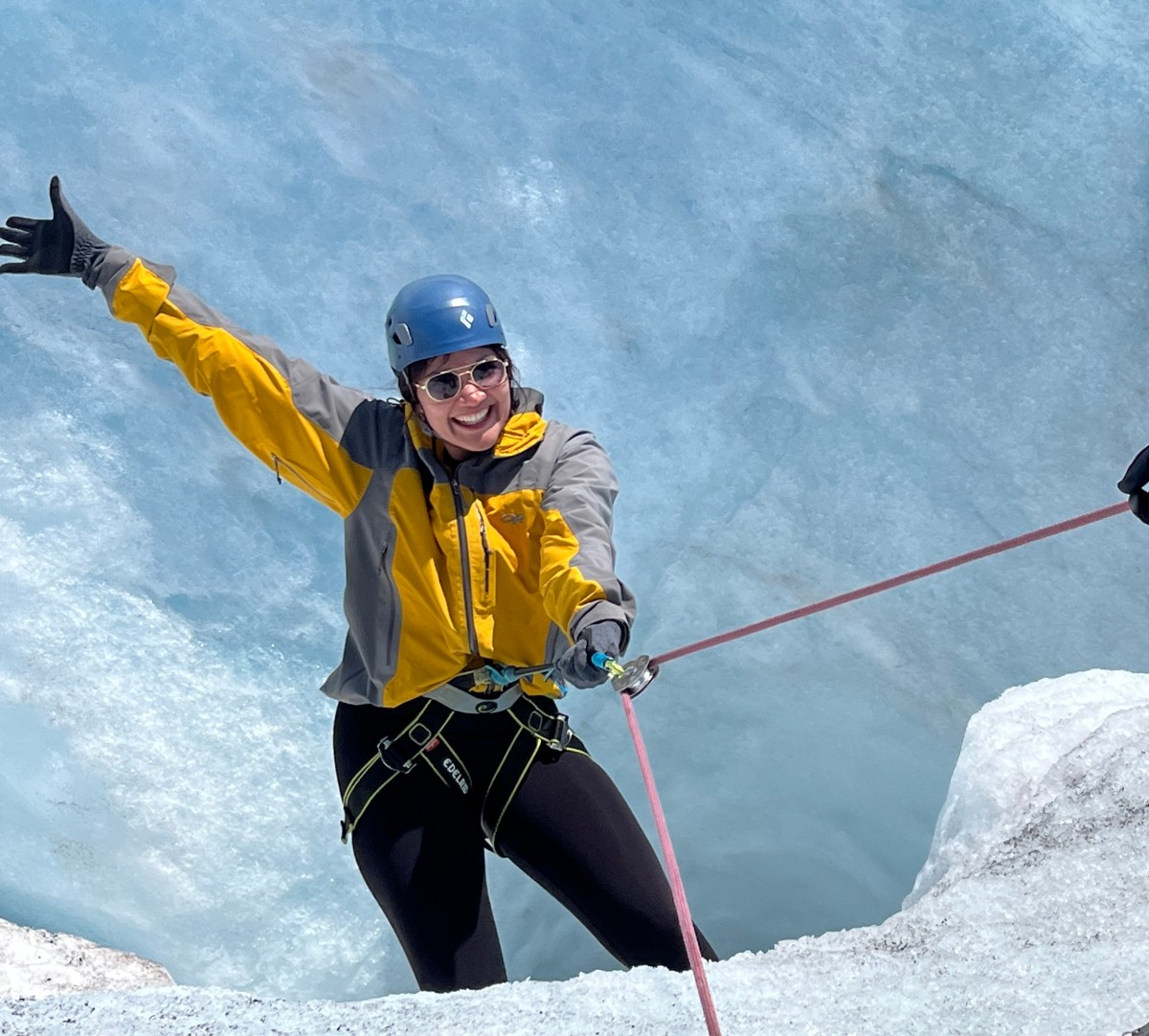 Person in yellow jacket and helmet smiling, climbing on icy surface with rope.